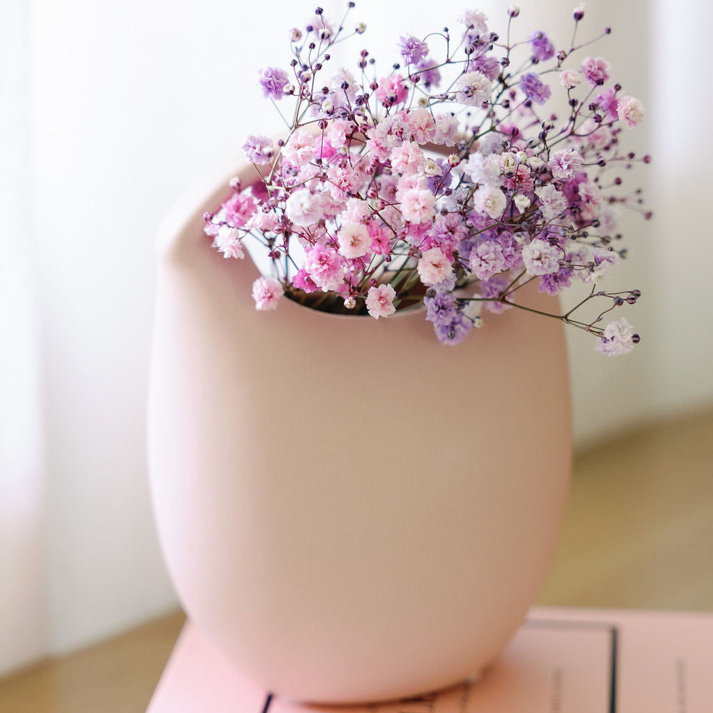Blush pink ceramic basket bud vase holding pink and purple baby's breath, styled on stacked fashion books.