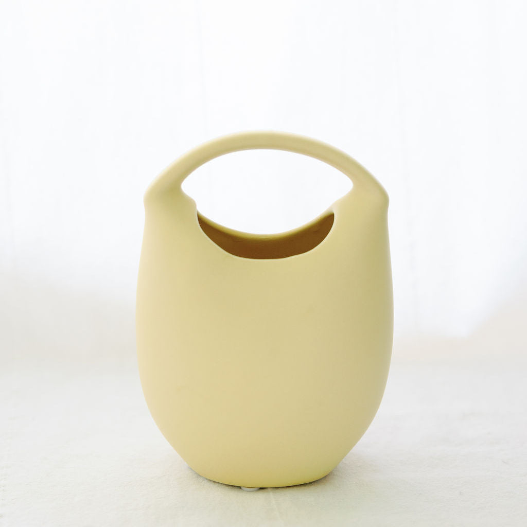Soft yellow ceramic basket bud vase with arched handle and scooped opening, shown empty on white.