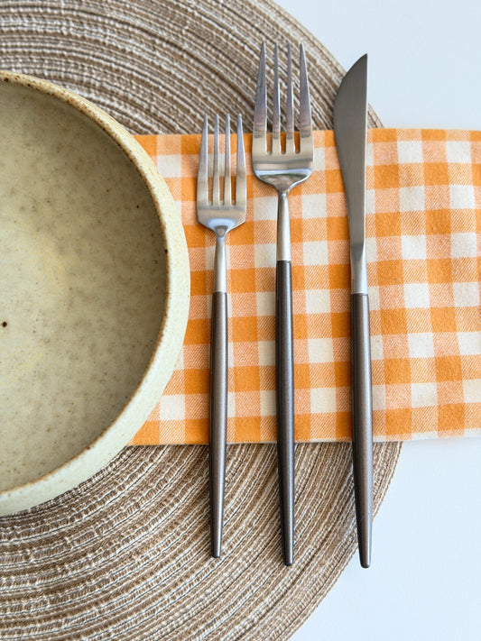 Matte stainless steel fork, fork and knife on orange gingham napkin beside a speckled bowl.