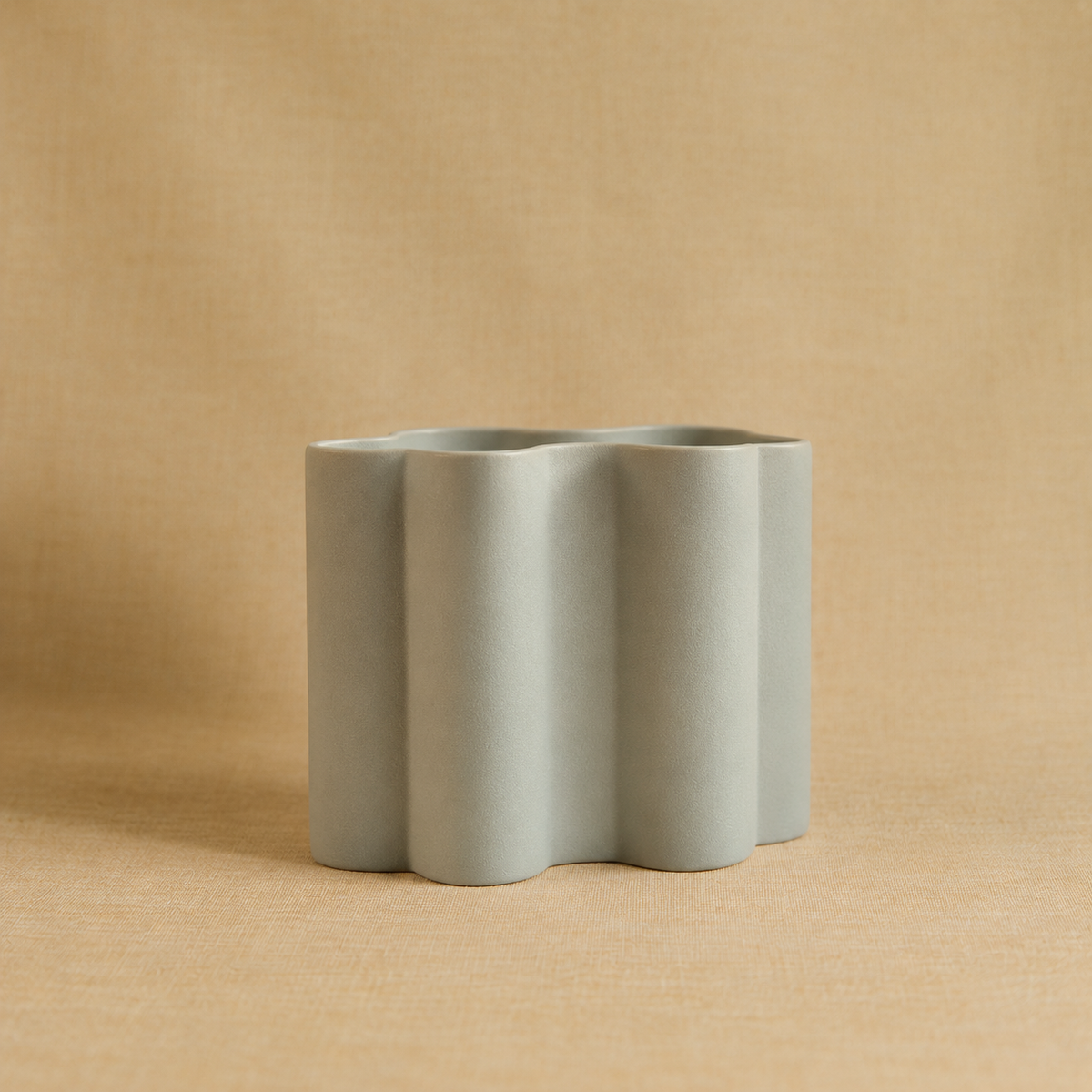 A modern, minimalist ceramic vase with a wavy, scalloped edge design in a muted sage green color, set against a warm beige textured background.