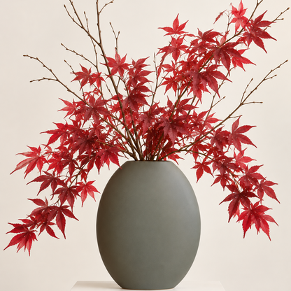 A matte dark gray ceramic vase filled with red maple leaves and brown branches, set against a solid light beige background.