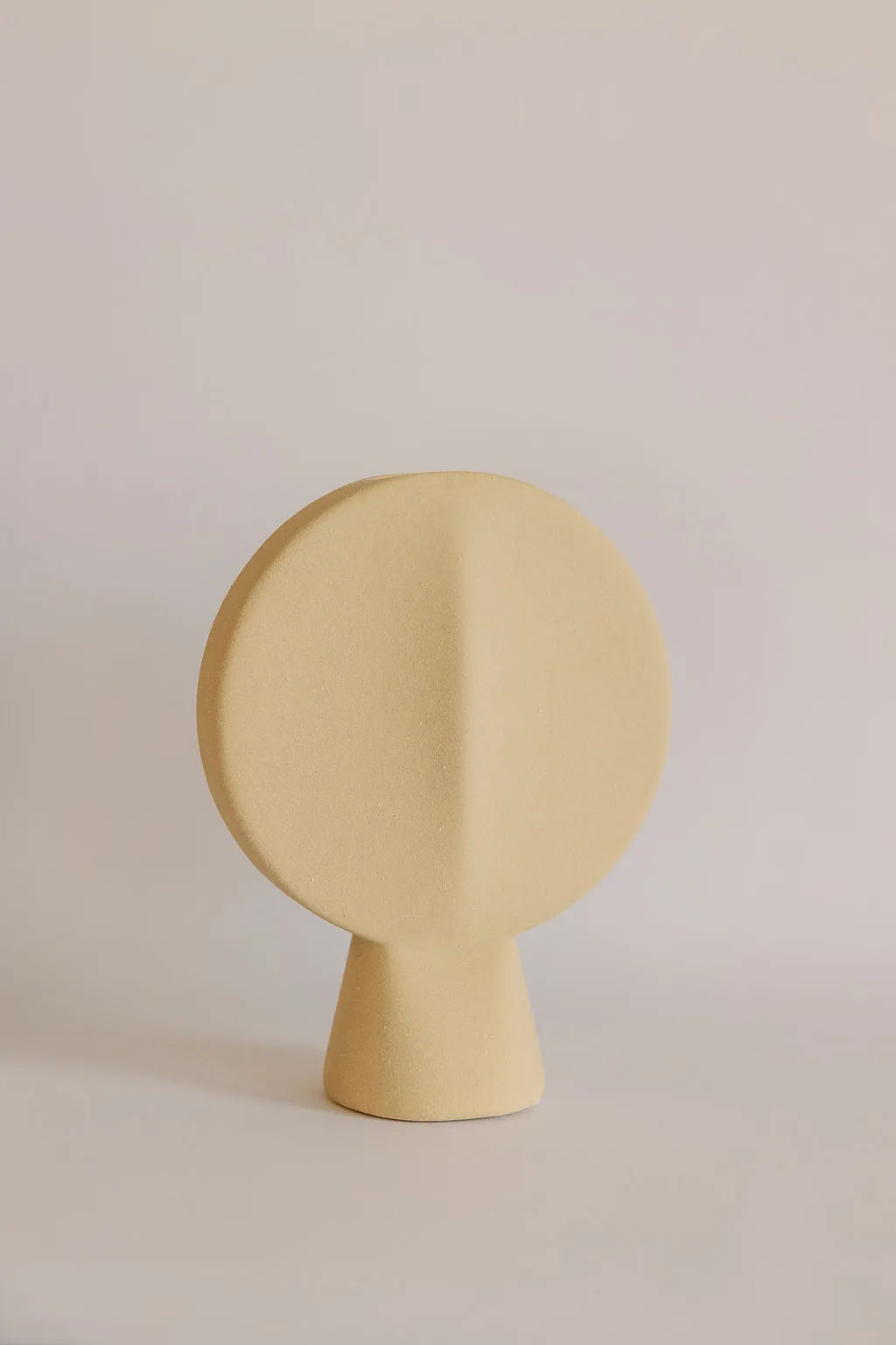 Beige ceramic bud vase shaped like a flat round disc on a tapered base, sandy texture