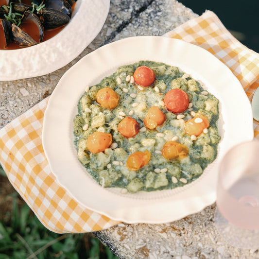 Provence Pleated scalloped white plate holding gnocchi with cherry tomatoes on a yellow gingham napkin outdoors.