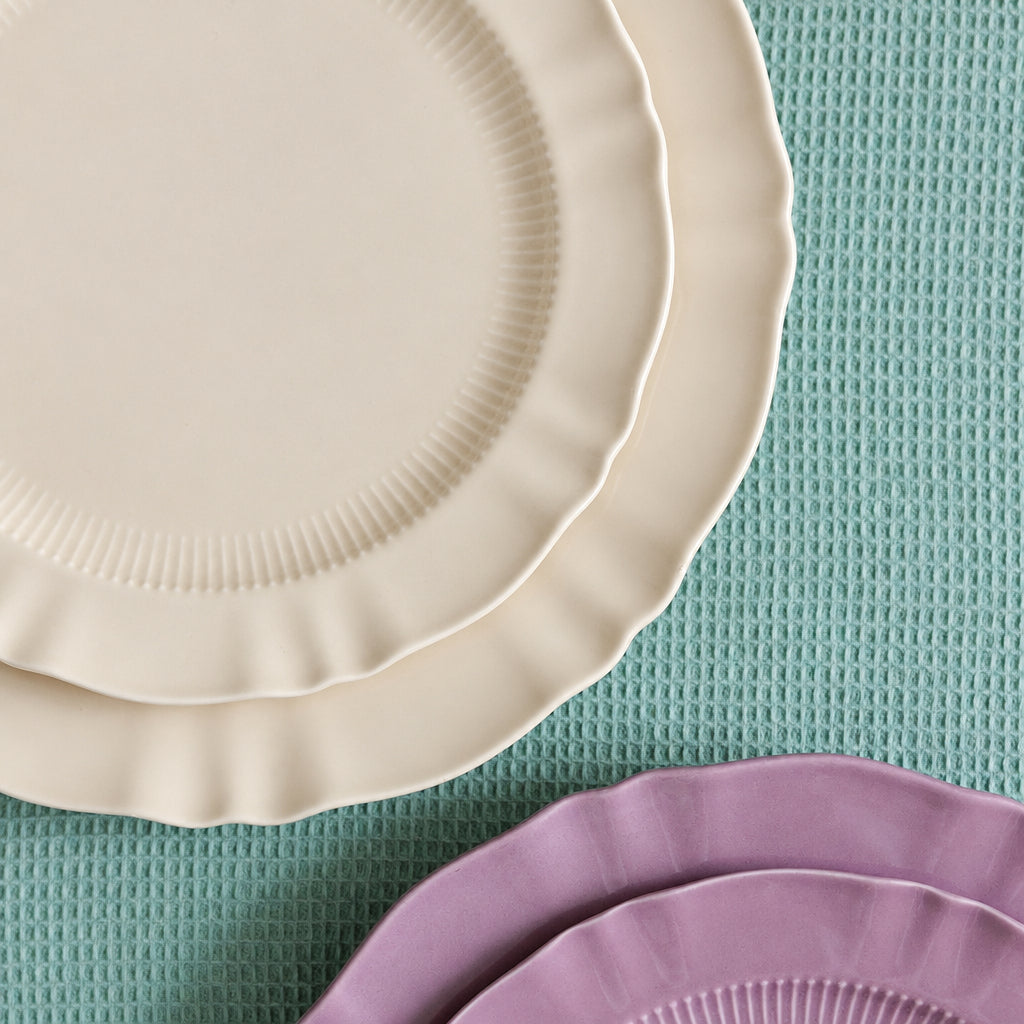 Close-up of scalloped ribbed rims on cream and mauve Provence Pleated porcelain plates over green fabric.