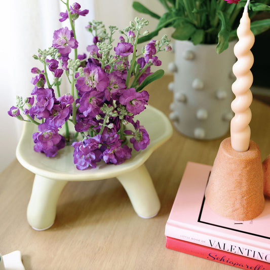 Cream Pedestal Bowl Vase on footed legs holding purple stock flowers on a wood table beside a candle holder.