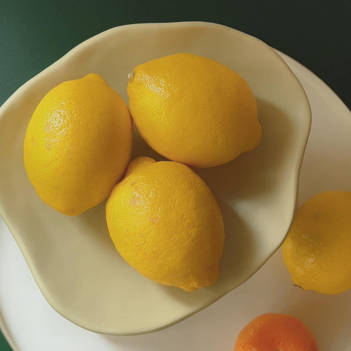 Wavy cream Pedestal Bowl Vase styled as a fruit bowl holding lemons, on a white plate atop a green table.