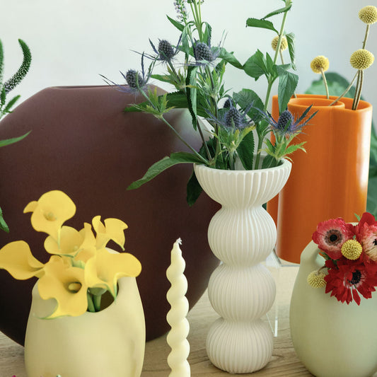 White ribbed Pleated Column Vase among colorful vases and a twist candle in a styled tabletop scene.