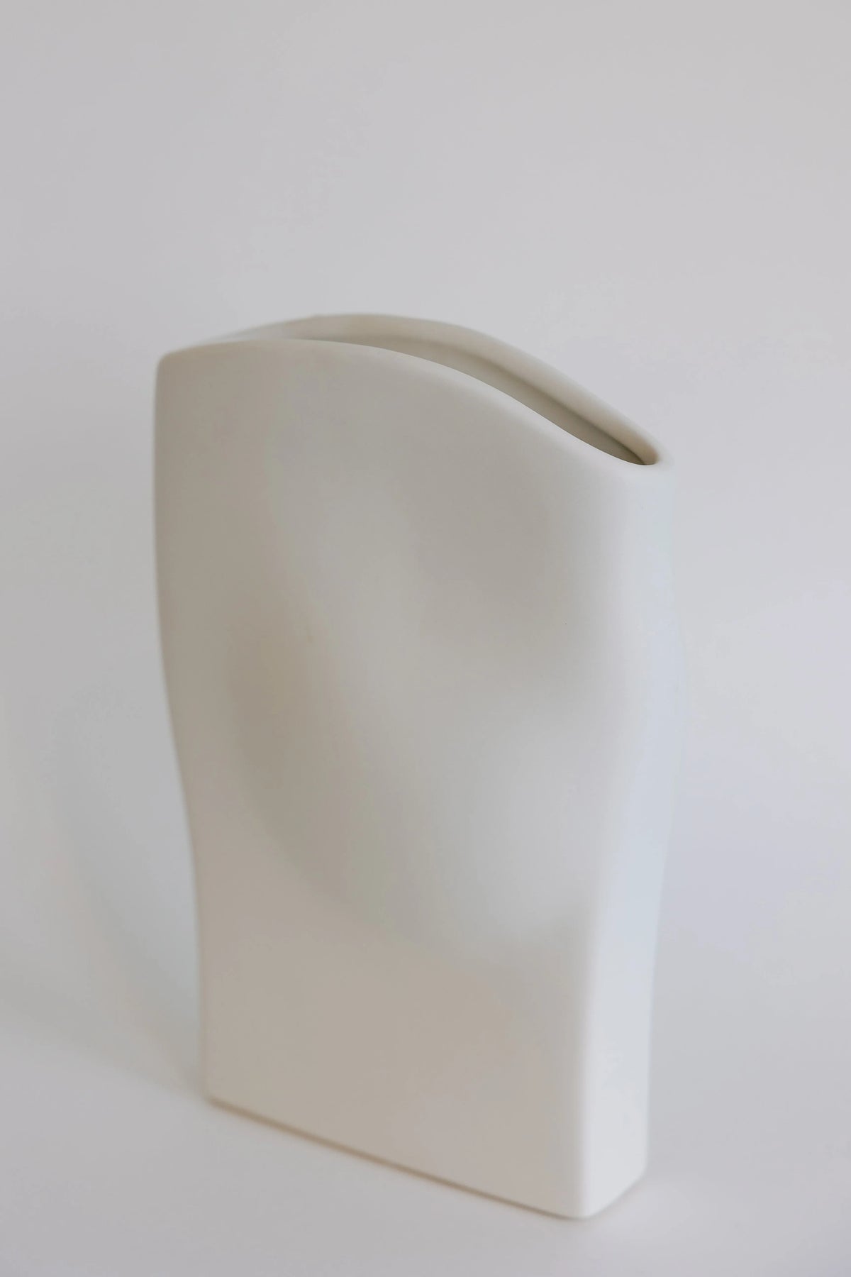 Wave ceramic vase in cream shown at an angle, flat draped body with sloping rim opening