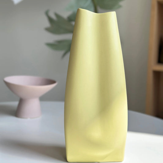 Bifold ceramic vase in soft yellow styled on a table, tall folded form with twisted body