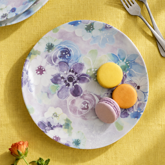 A round ceramic plate with a watercolor floral pattern in purple, blue, and white, styled on a textured yellow tablecloth with macarons and cutlery.
